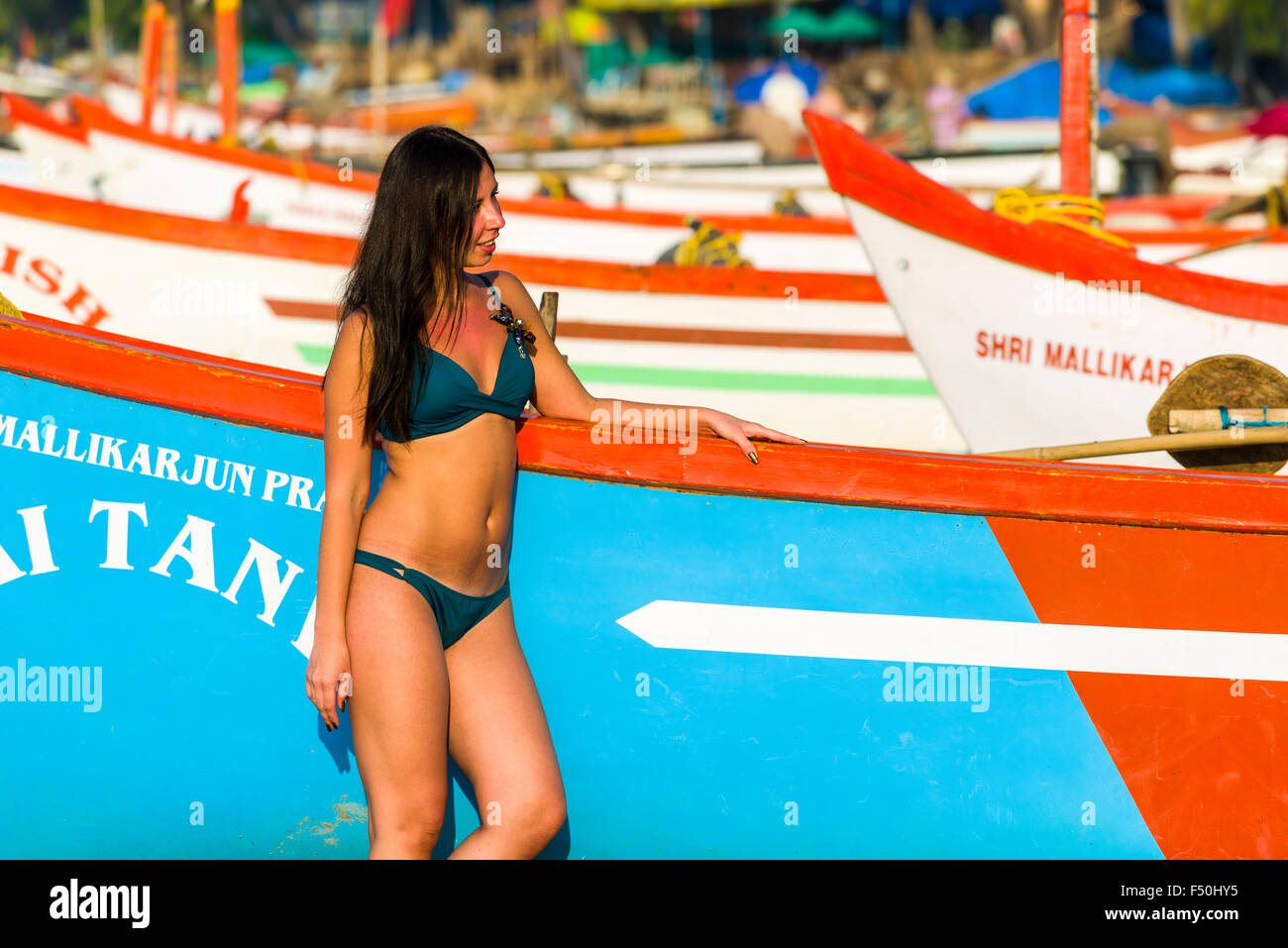 A young woman, wearing bikini, is posing for photographs in front of a fishing boat at Palolem Beach Stock Photo
