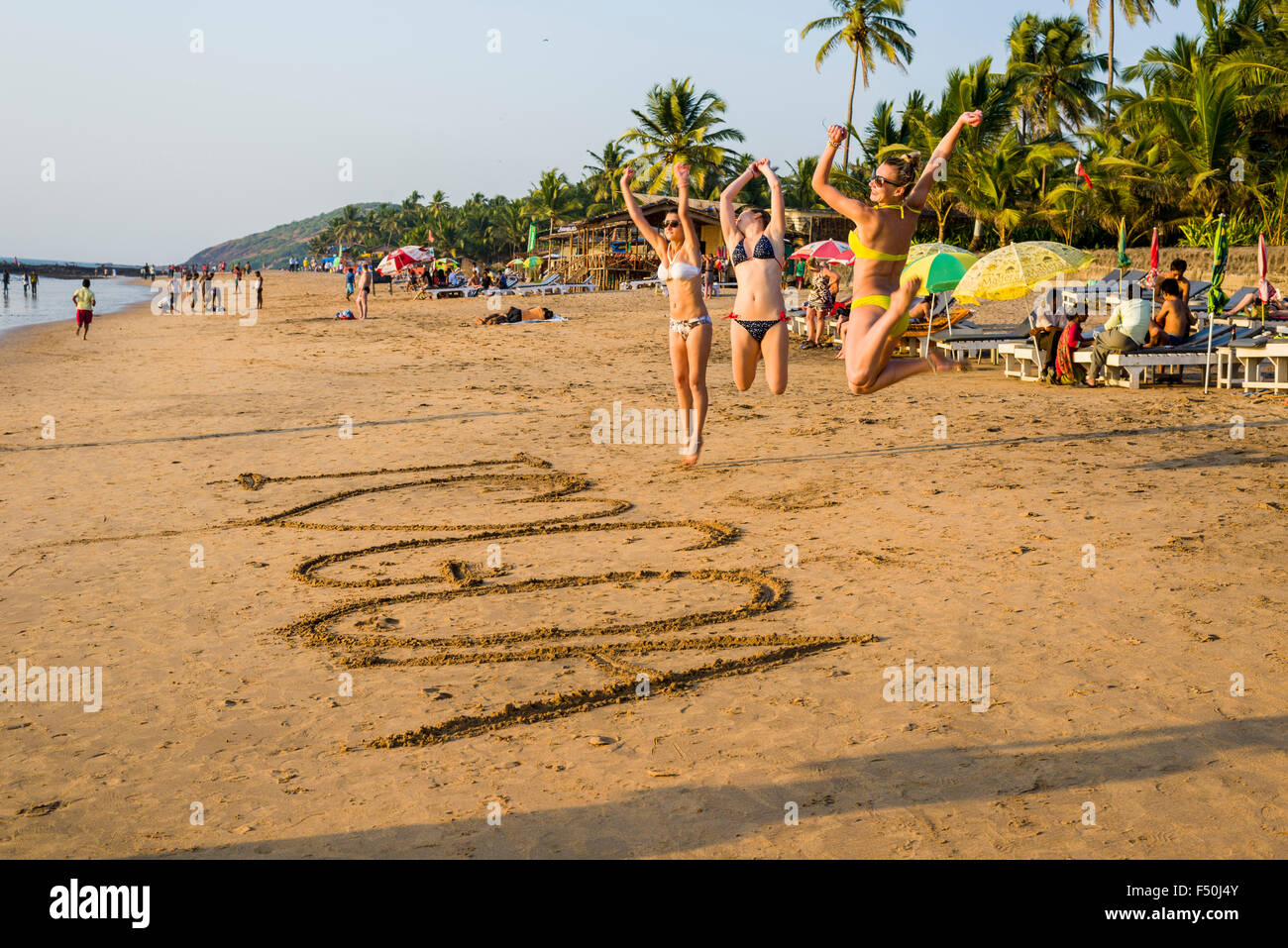 Three young women wearing bikinis are jumping after writing 'I love Goa' in the sand of Anjuna Beach Stock Photo