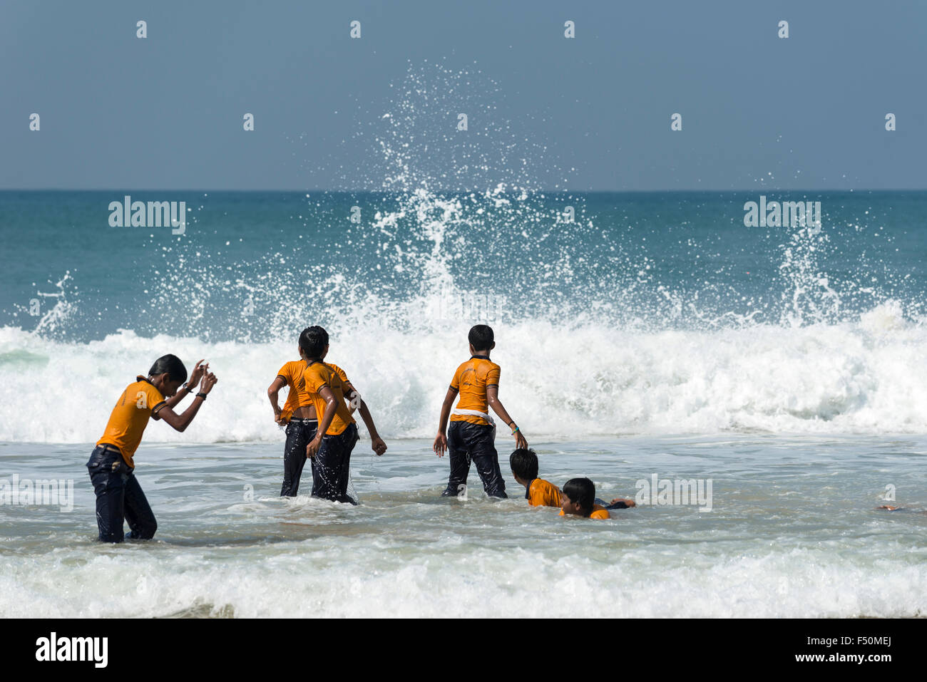 A group of schoolboys, wearing yellow shirts, is standing on the beach, some are playing in the waves Stock Photo