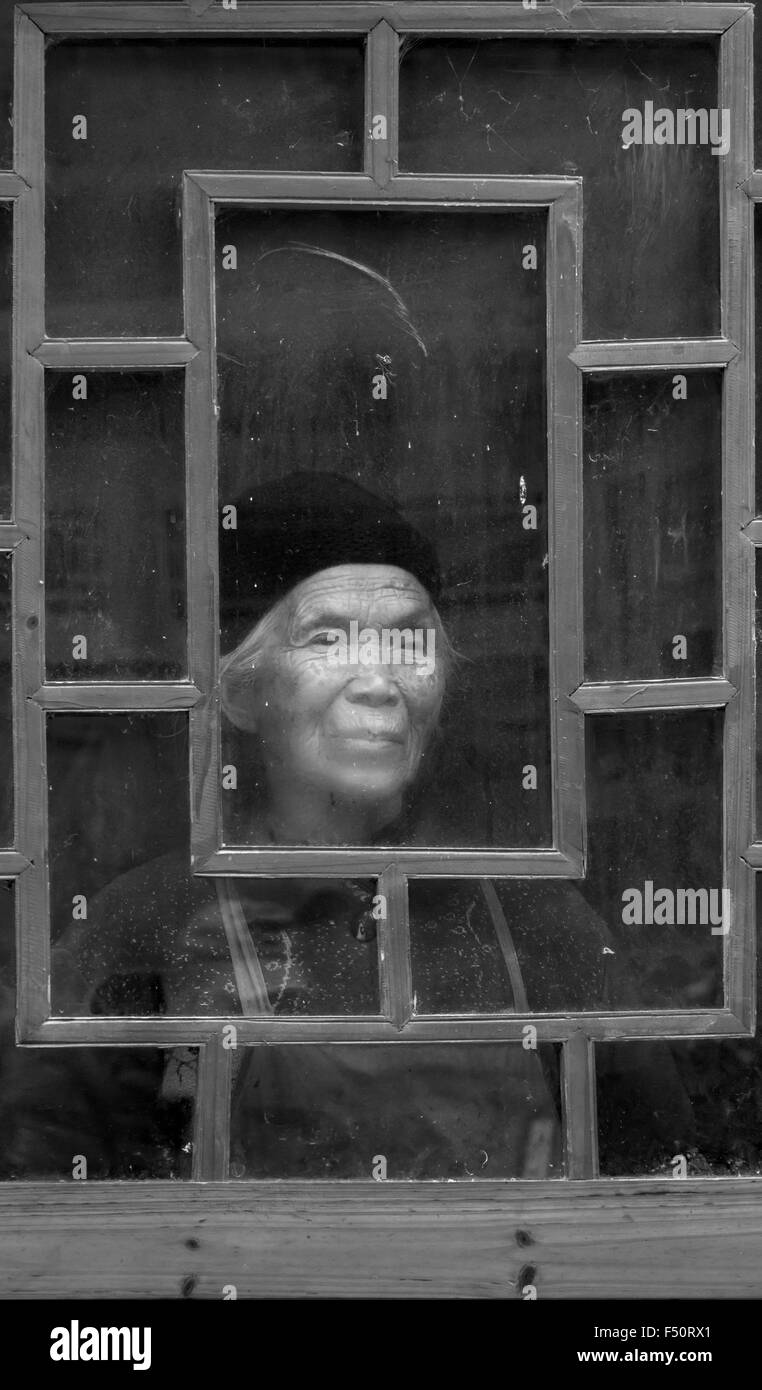 Old woman at a dirty window on a cold day BW, Shiqiao Miao Village, Guizhou Province, China Stock Photo