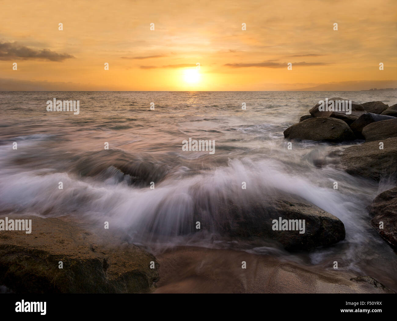 Ocean sunset is waves rushing over sand on rocks along the beach as the sun sets in a colorful soft cloud filled sky in the back Stock Photo