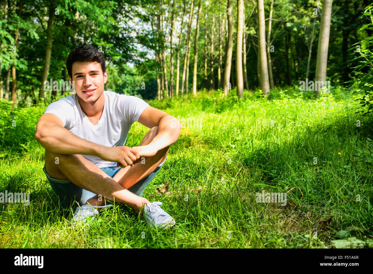 Attractive, fit young man relaxing sitting on lawn grass in the countryside among trees, looking at camera, smiling Stock Photo