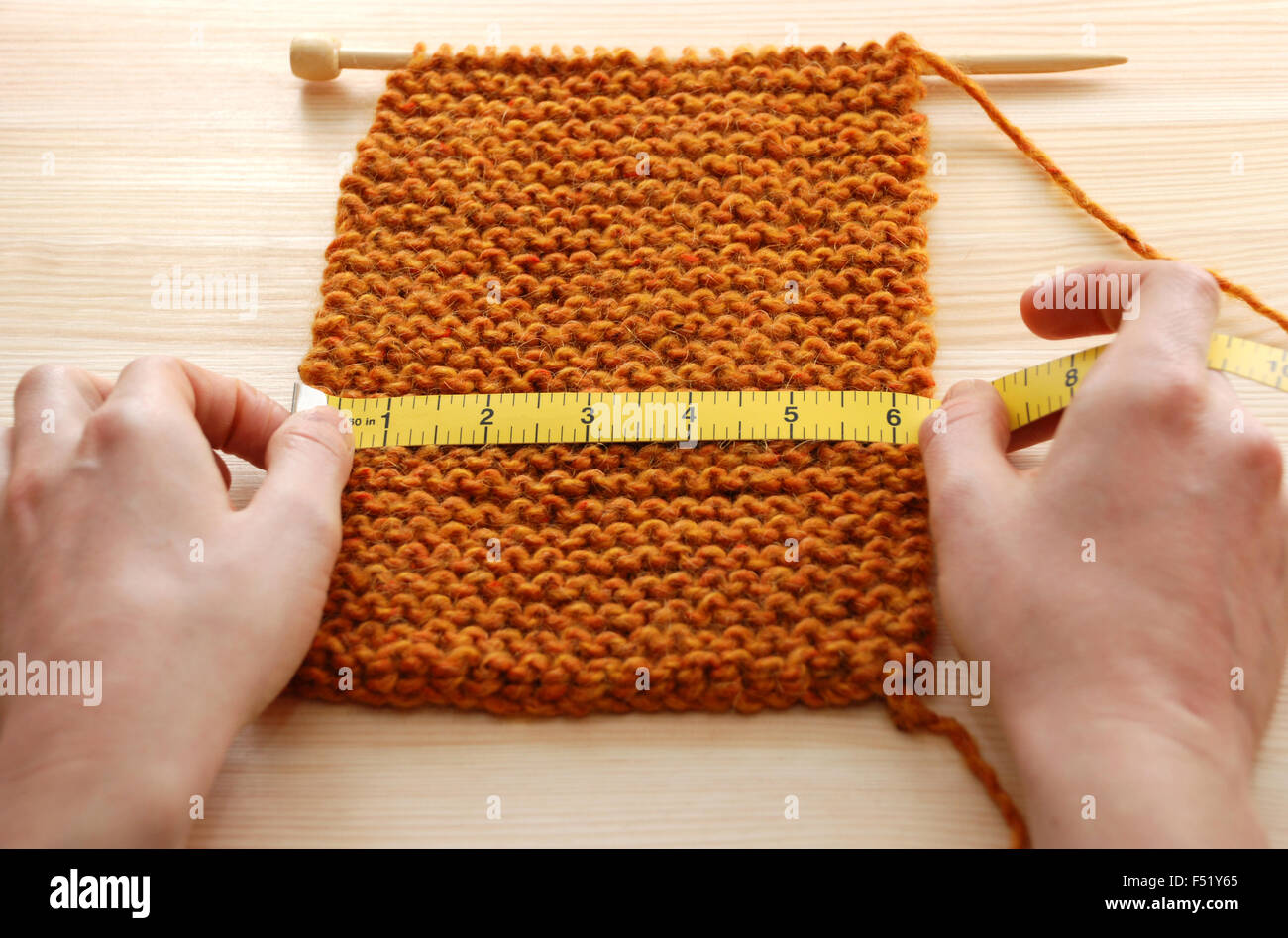 Two hands hold a tape measure across a piece of knitting on a wooden table, measuring in inches Stock Photo