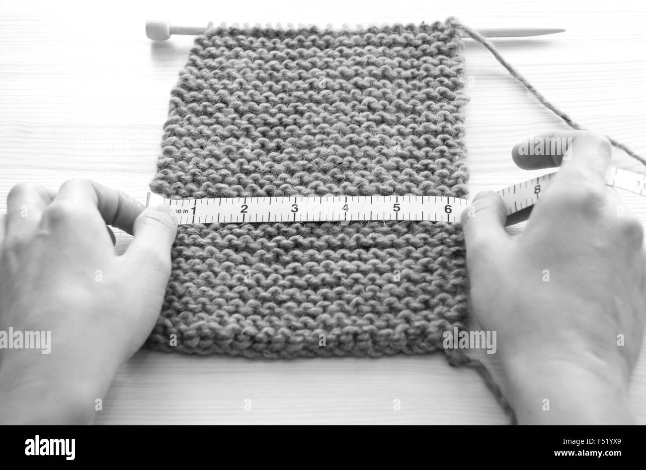 Two hands hold a tape measure across a piece of knitting on a wooden table, measuring in inches - monochrome processing Stock Photo