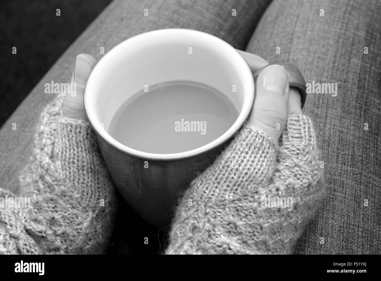 A woman in a warm jumper holding a hot cup of tea or coffee on her lap - monochrome processing Stock Photo
