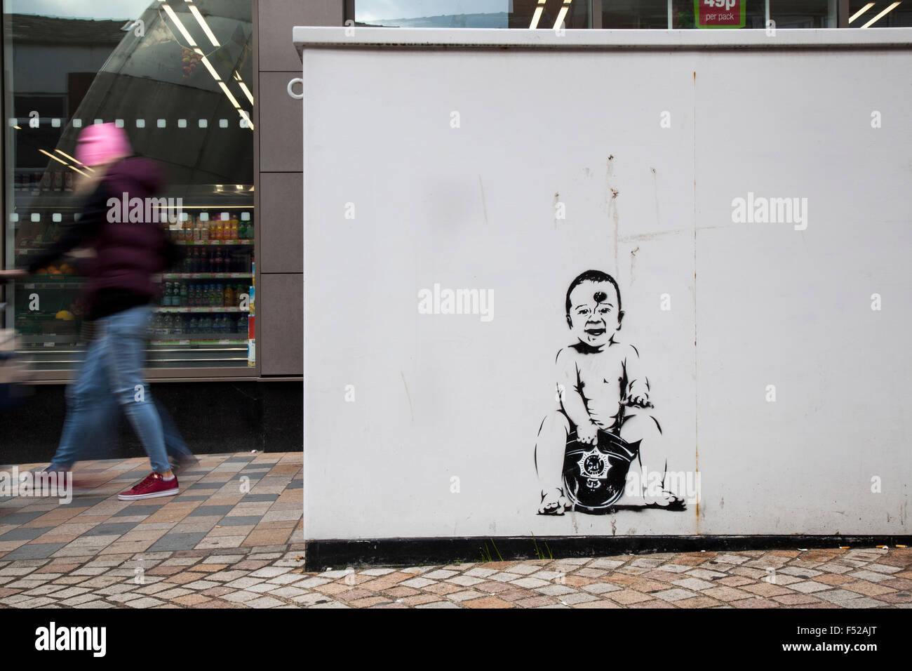 Pedestrian walking past street art, graffiti wall painting, decoration. Baby sitting on Policeman's Helmet; Banksy style artwork in Birley street, UK Stock Photo