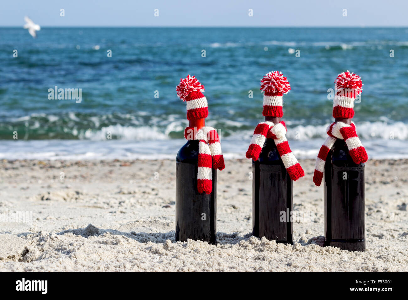 Christmas gifts. Three bottles of wine in knitted hats and scarves on the beach. Selective focus. Stock Photo