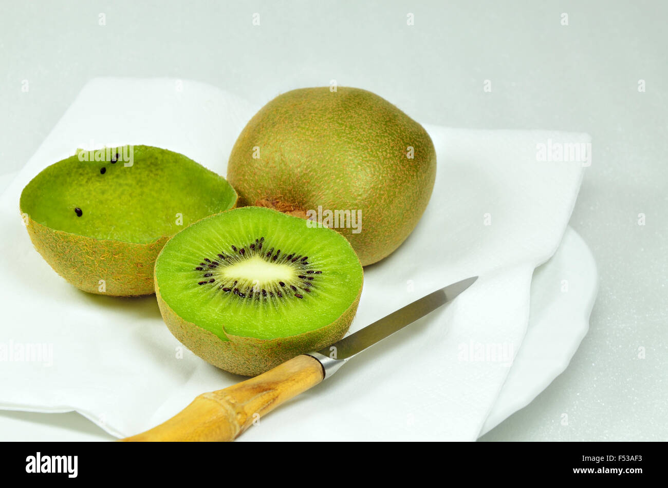 three kiwi fruits, halved and whole, full and empty, on white plate with knife, close up, macro