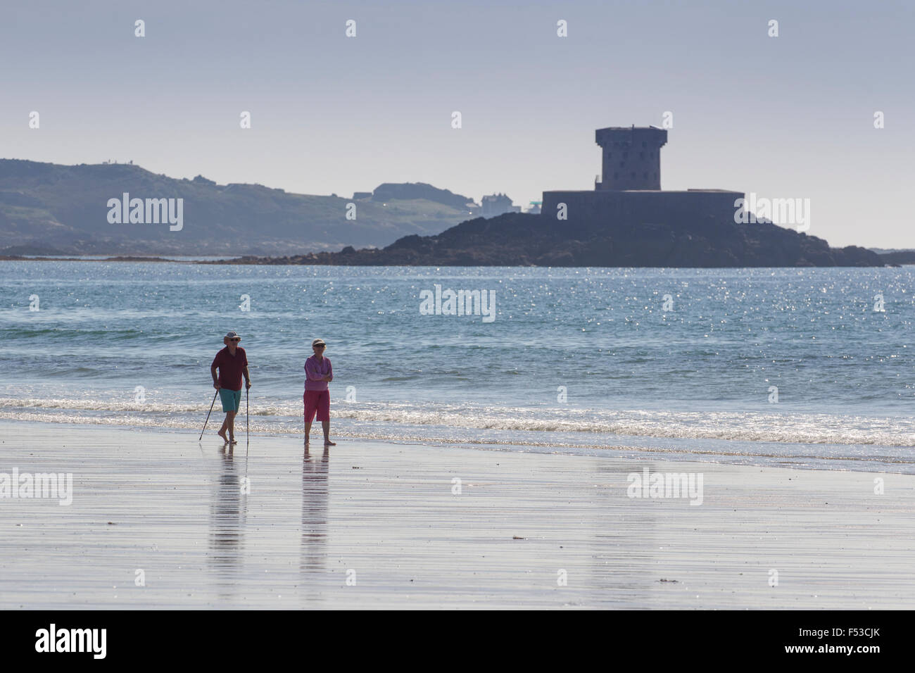 Pensioner couple walking along the shore line on St St Ouen's beach with La Rocco Tower in the background Jersey Channel Islands Stock Photo