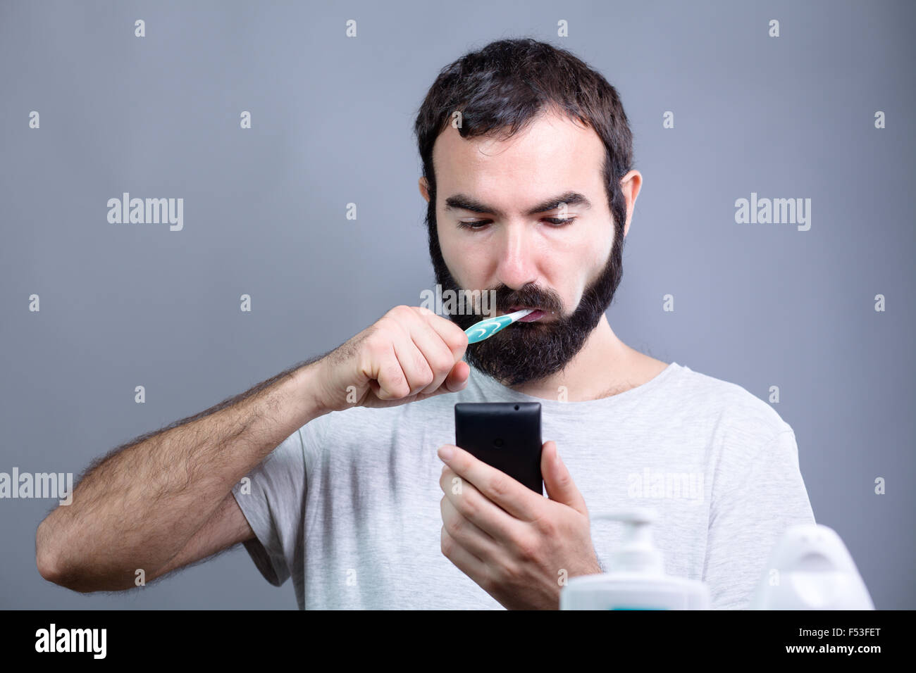 Man with Beard Washing His Teeth with a Toothbrush while Watching a Smartphone Stock Photo