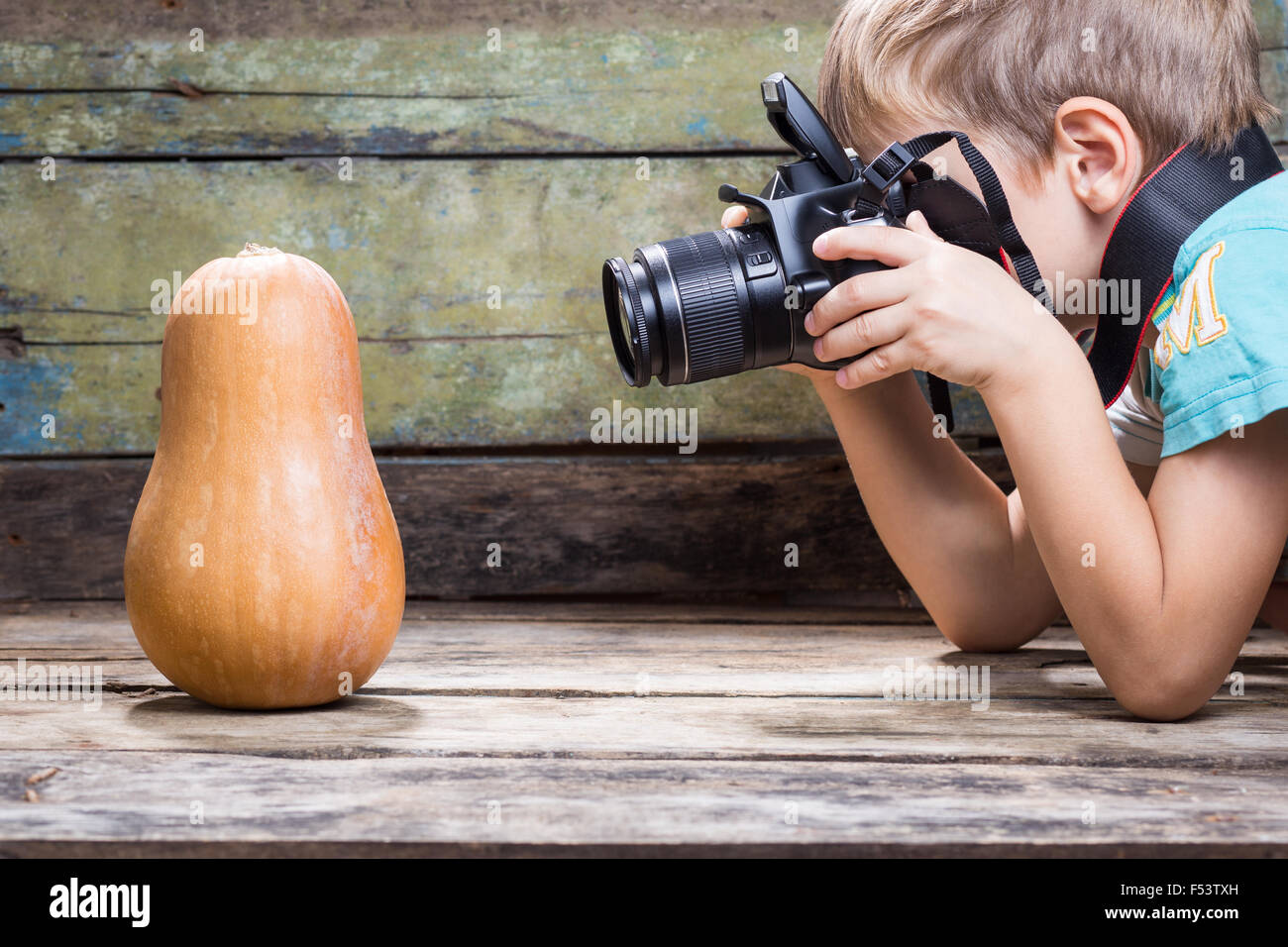 Funny fair boy use DSLR camera trying to take photo of ripe pumpkin on wooden background in studio. Photography lessons or learn Stock Photo