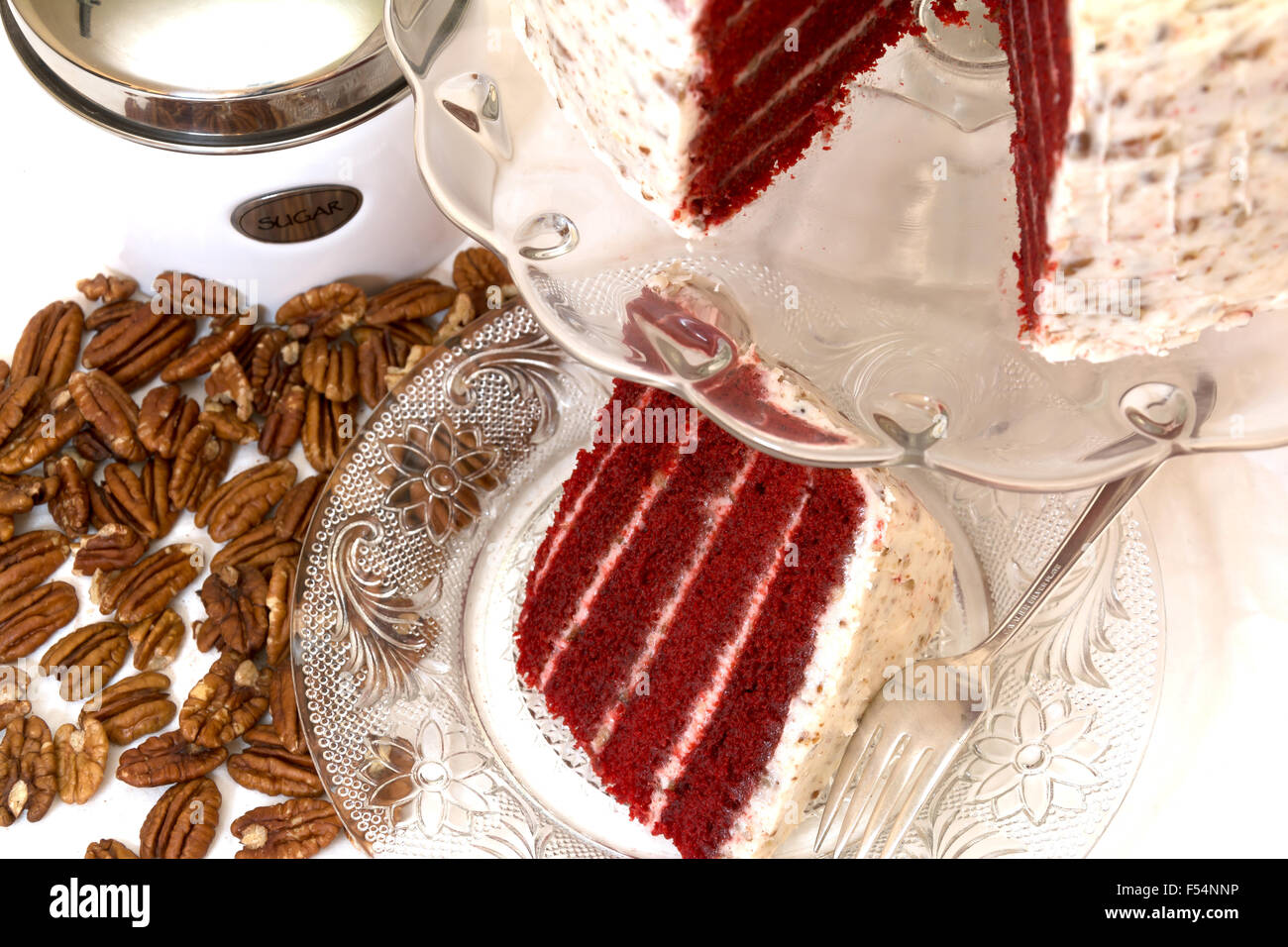 Slice of red velvet cake and pecans with sugar canister in background.  Slice is removed from whole cake which is in background. Stock Photo