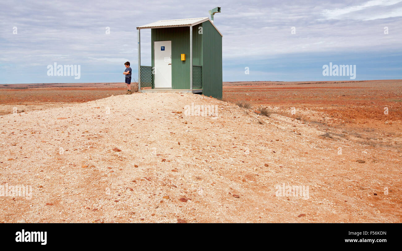 Humourous view of young boy beside isolated public toilet on vast barren red outback plains in remote area of Australia Stock Photo