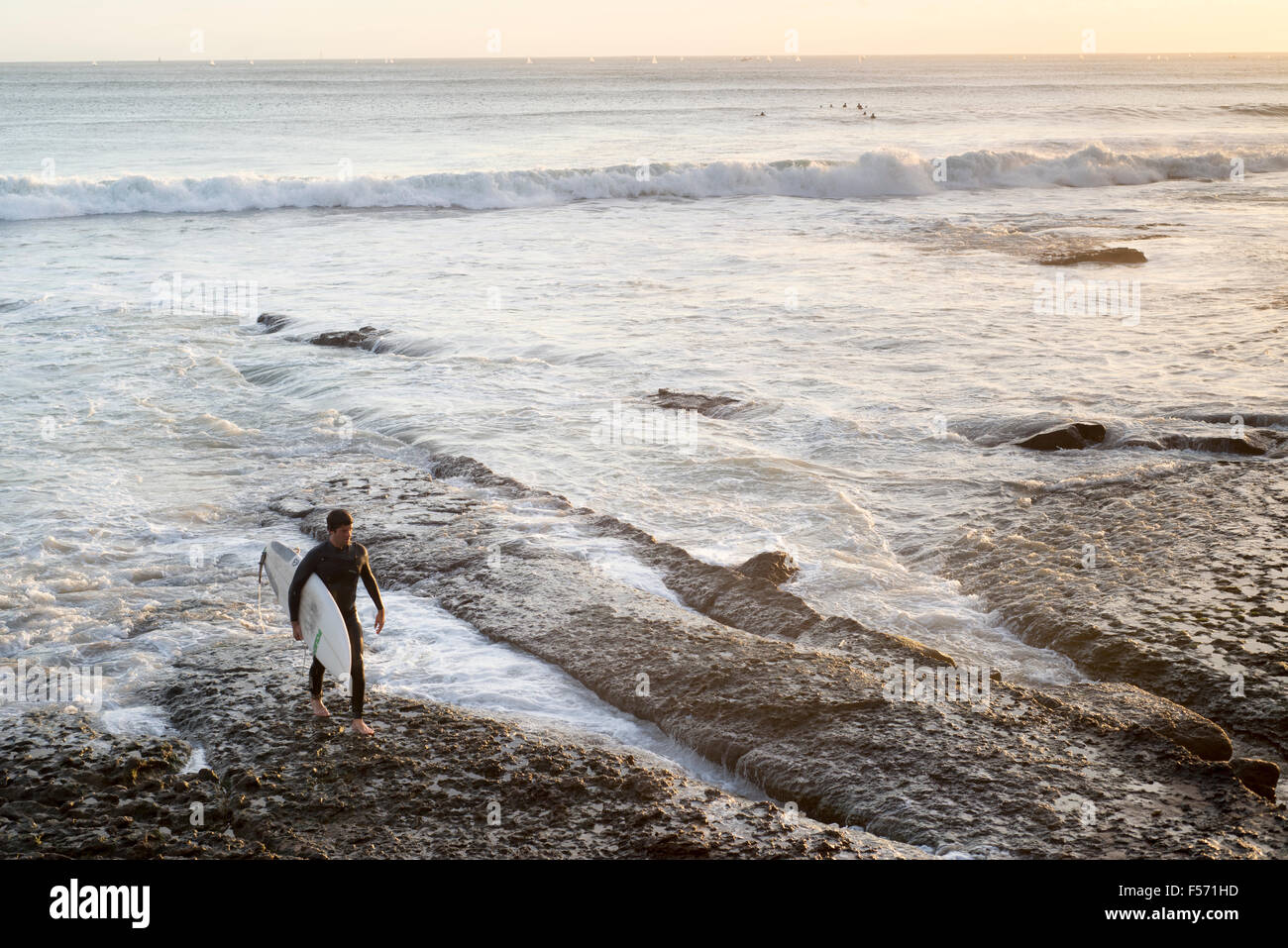Male surfer walking over reef looking out to sea at surf with setting sun, Estoril, Cascais, Portugal Stock Photo