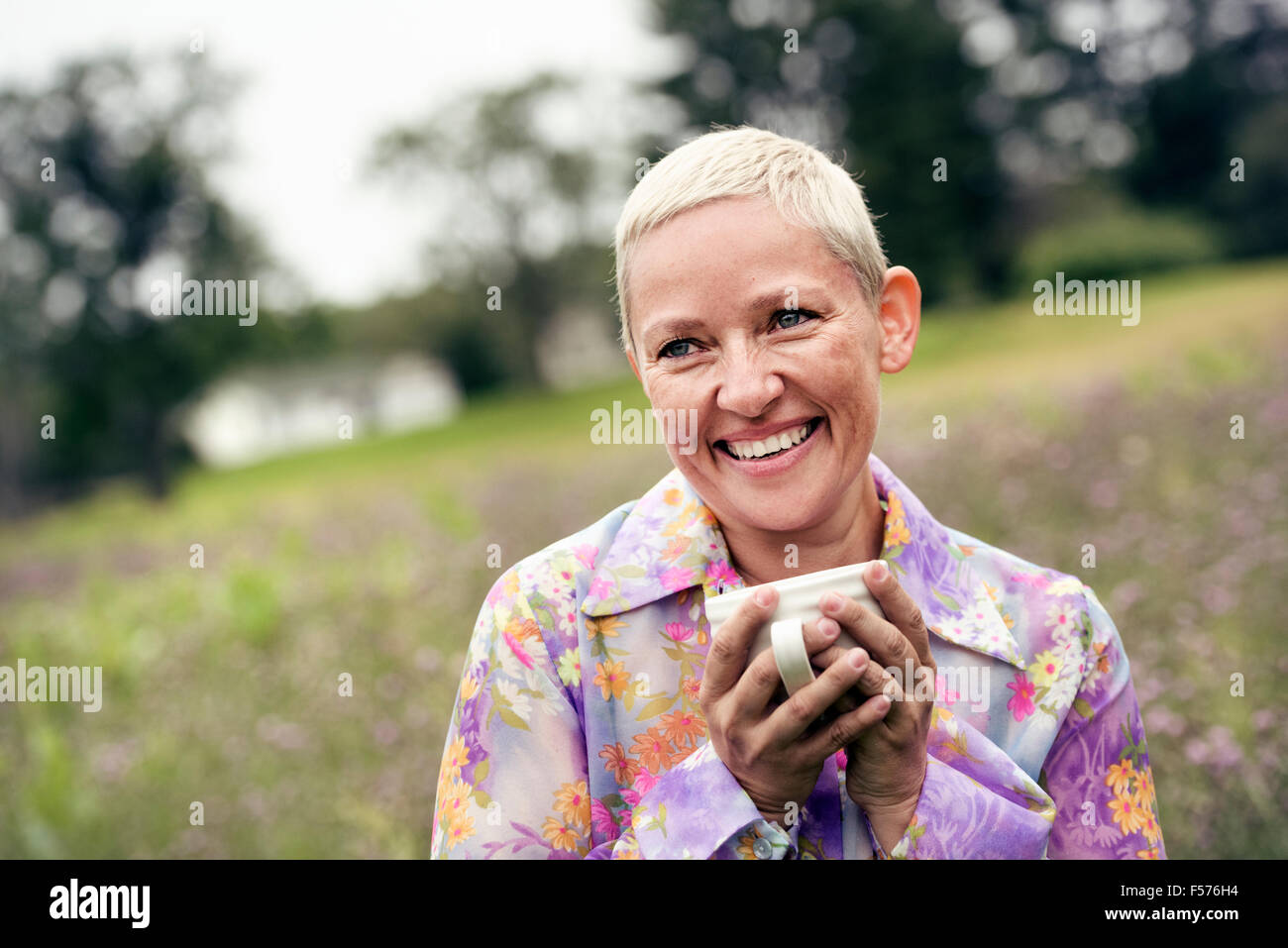 A woman with her hands wrapped around a coffee cup, sitting on a meadow. Stock Photo