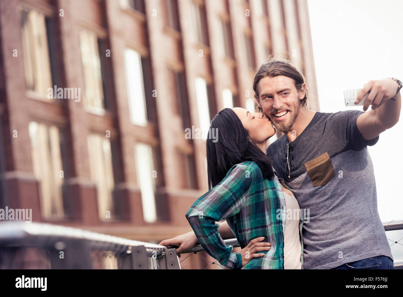 A woman kissing a man on the cheek, posing for a selfie by a large city building Stock Photo