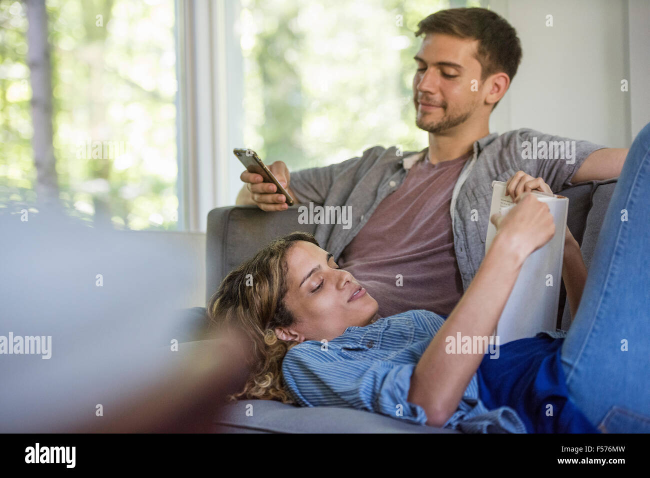 A couple, a man checking his phone and a woman lying down reading a book. Stock Photo