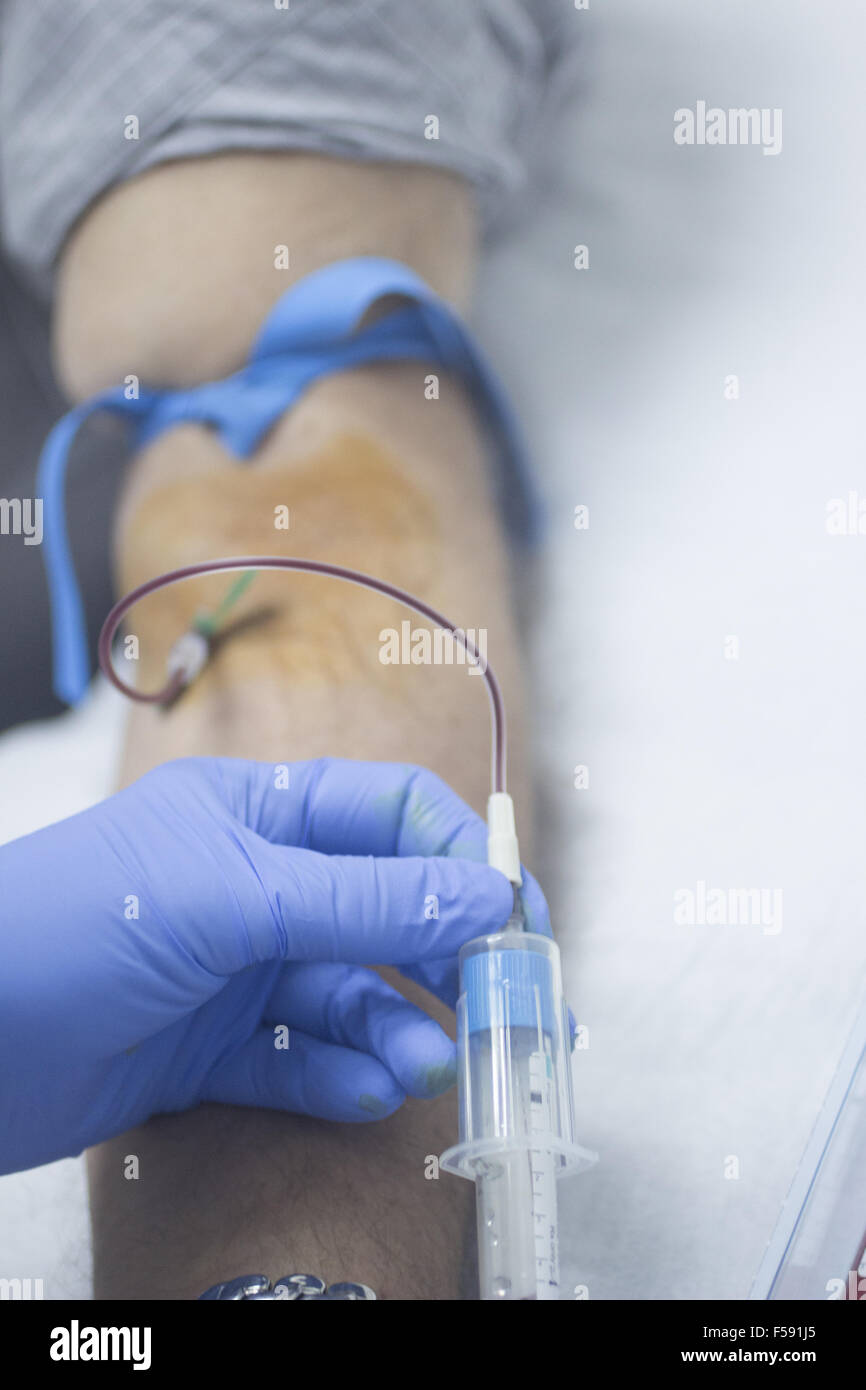 Close-up color photo of a female hospital clinic nurse wearing sterile blue gloves and senior male patient aged 65-70 giving a blood donation sample from the interior of his elbow resting arm on sterile blue white defocused background. Stock Photo