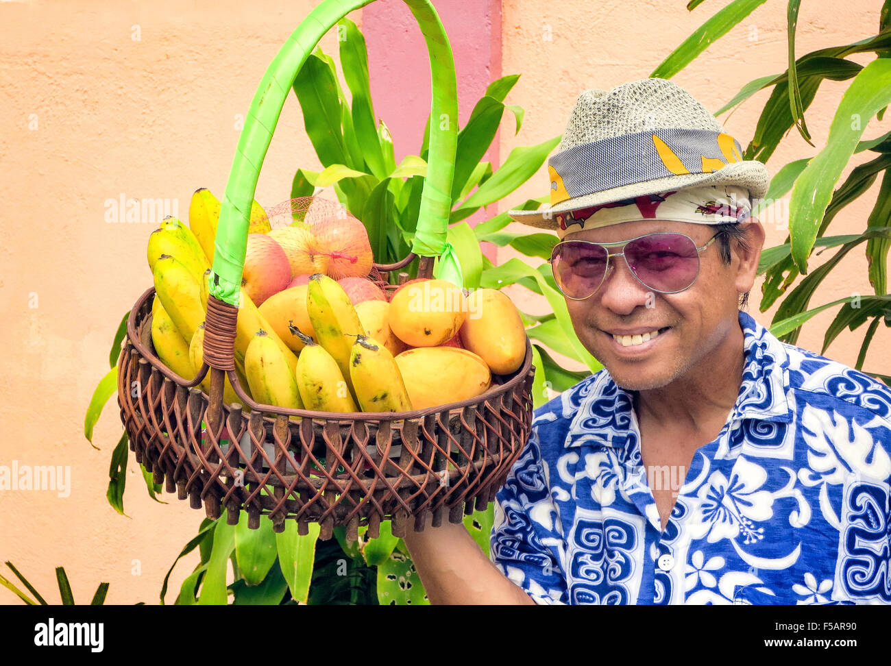 Portrait of a smiling Filipino man wearing a fedora hat, sunglasses and a flower shirt holding a basket of fresh fruit. Stock Photo
