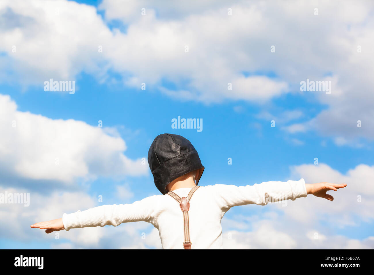 Little boy in vintage clothes dreams of flying with outstretched arms in front of the cloudy sky (copy space) Stock Photo