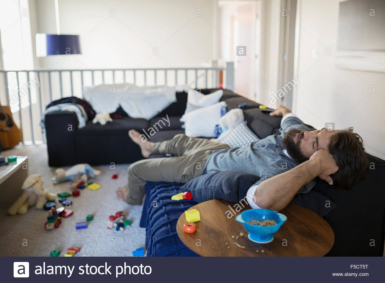 Exhausted man napping on sofa surrounded by toys Stock Photo