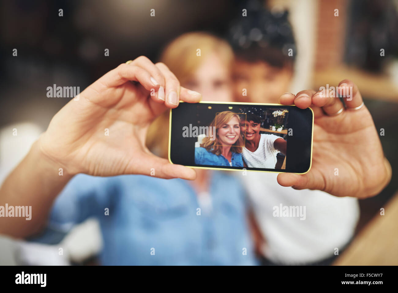 Two attractive young women friends taking a selfie on a mobile with the image displayed to the camera on the screen, young multi Stock Photo