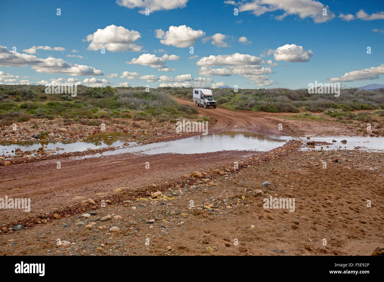 Four wheel drive campervan / motorhome about to drive through water at creek crossing on Australian outback road hemmed by low vegetation after rain Stock Photo