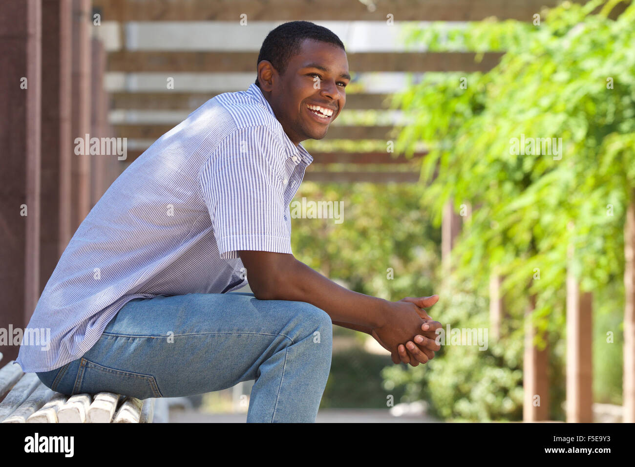 Close up portrait of a smiling relaxed young black guy sitting on bench ...