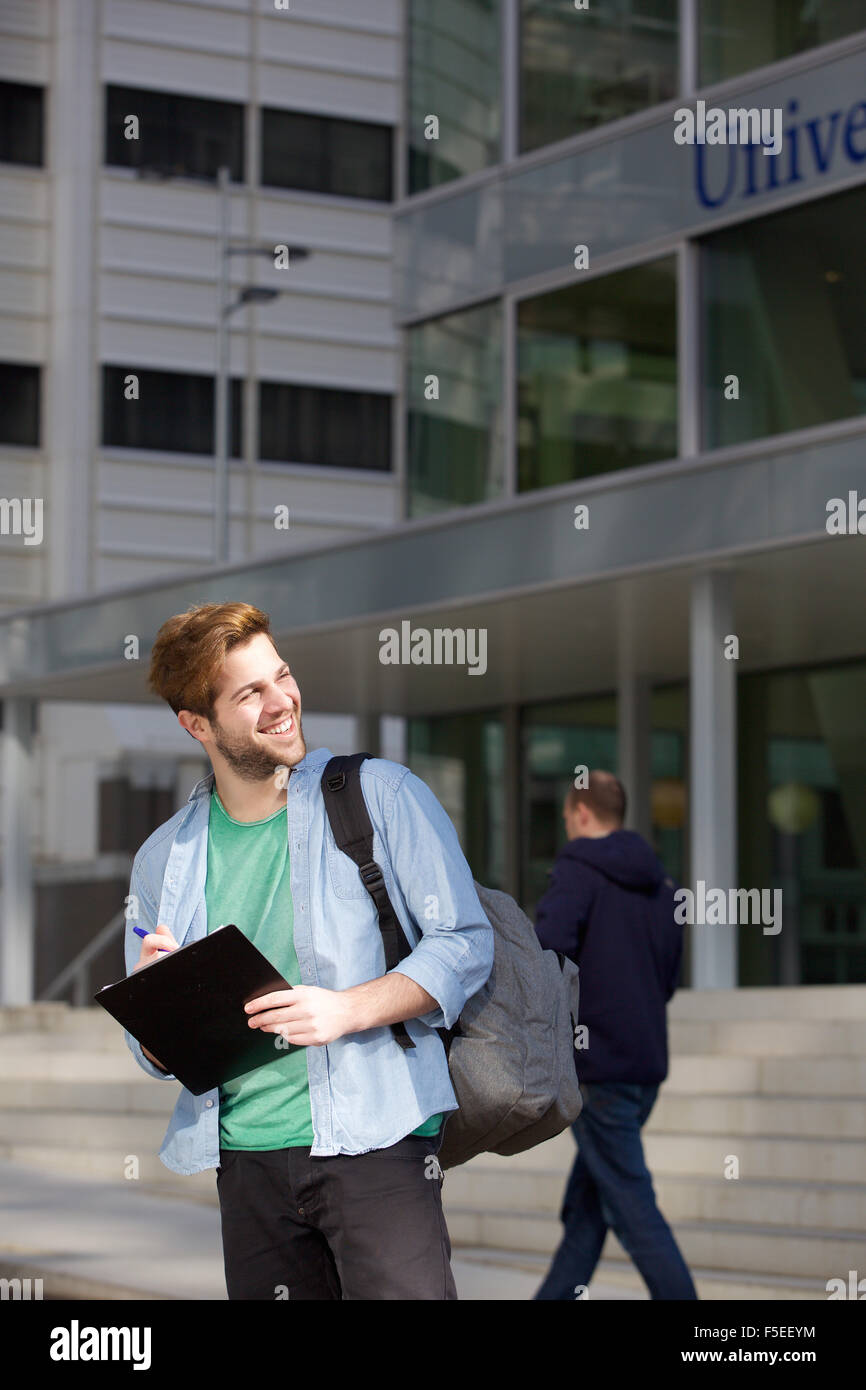 Portrait of a happy male university student standing on campus with pen ...