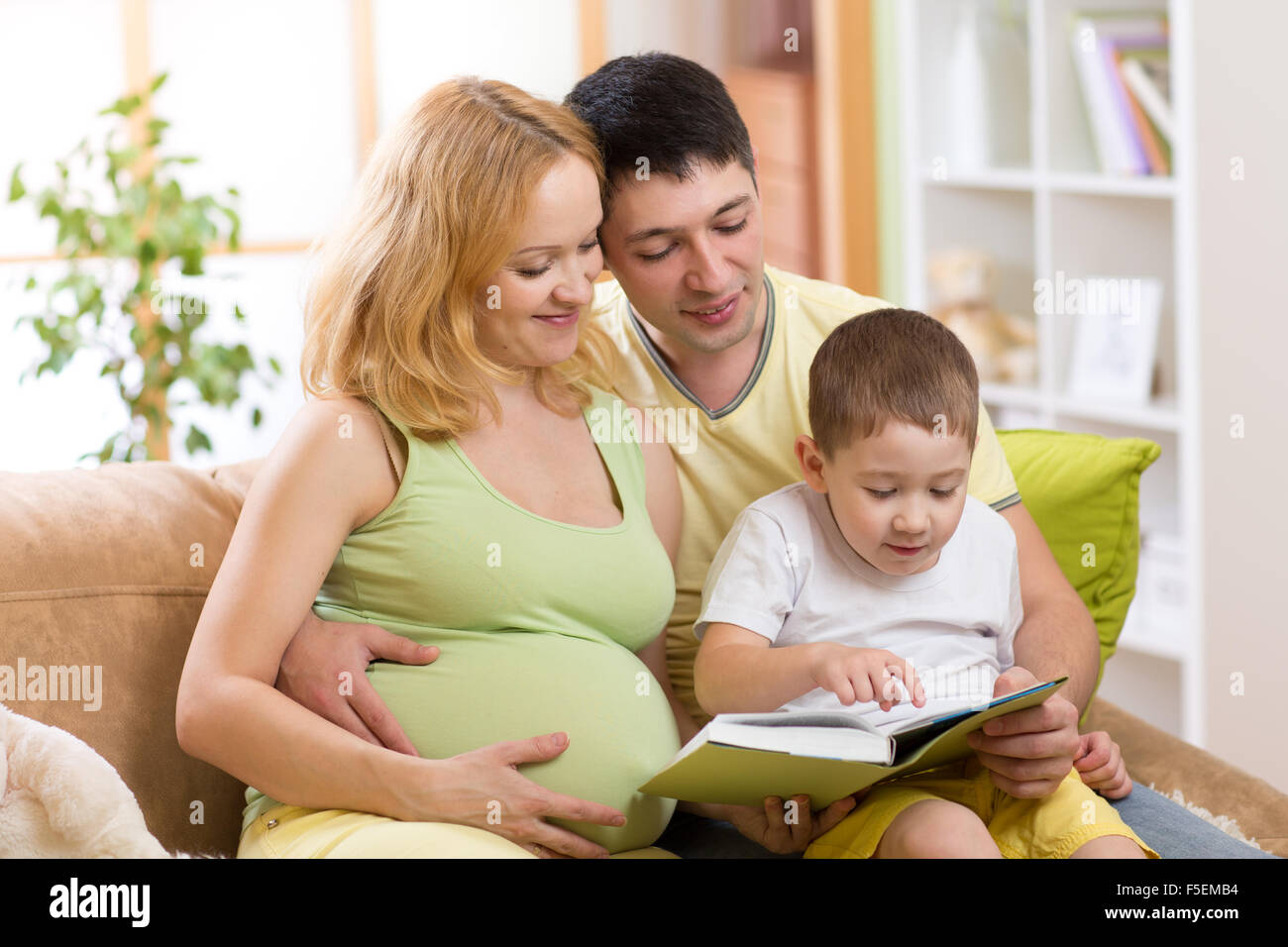 Happy parents reading book to child together on couch in home Stock Photo