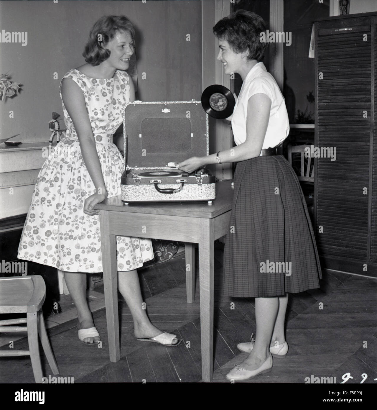 1950s historical picture of two young adult girls dressed in the clothes of the era, excited at playing an LP or vinyl record on a small portable record player. Stock Photo
