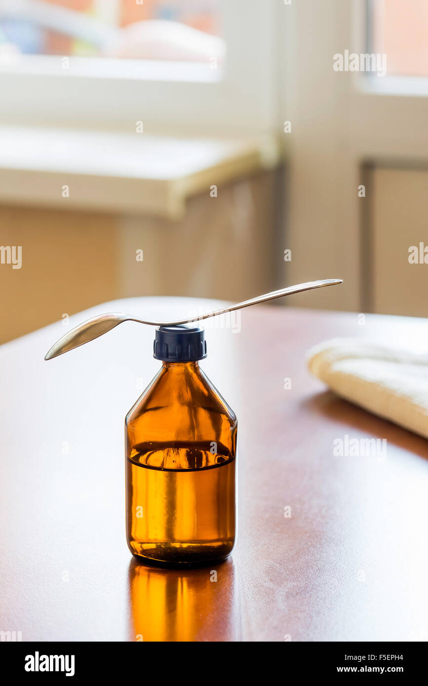 A cough syrup bottle with a spoon on the table close to the window
