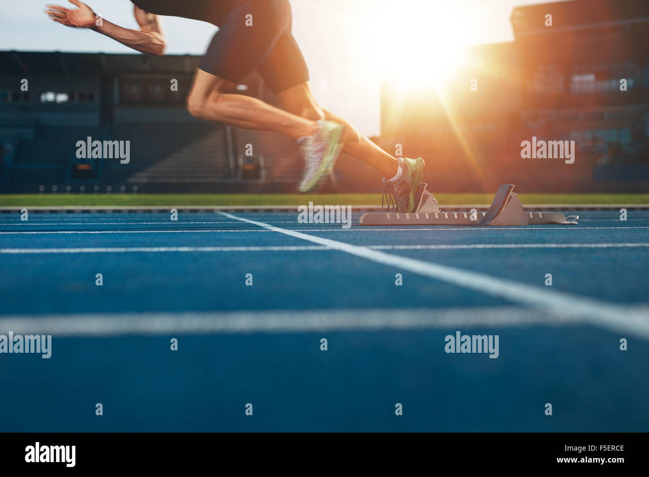 Athlete running on athletic racetrack. Low section shot of male runner starting the sprint from the starting line with bright su Stock Photo