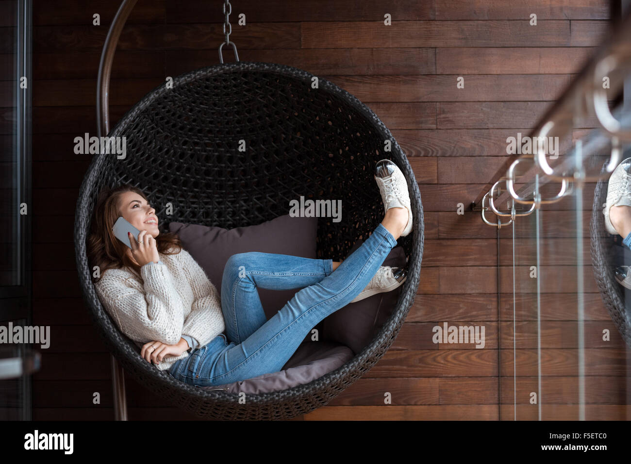 Beautiful happy girl weared in white sweater and blue jeans lying in a bubble chair and talking  on the cellphone Stock Photo