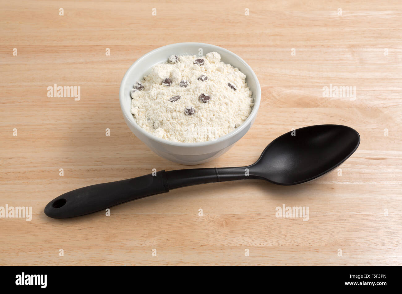 Dry chocolate chip cookie mix in an old bowl with a black handle spoon in the foreground atop a wood table top illuminated with Stock Photo