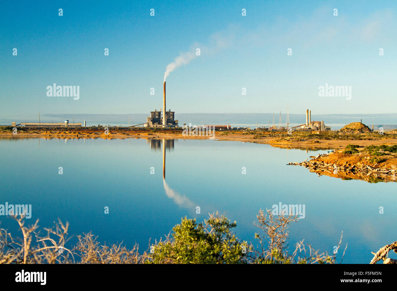 Coal fired power station with smoking chimney stack reflected in mirror surface of blue water of Spencer Gulf at Port Augusta SA Stock Photo
