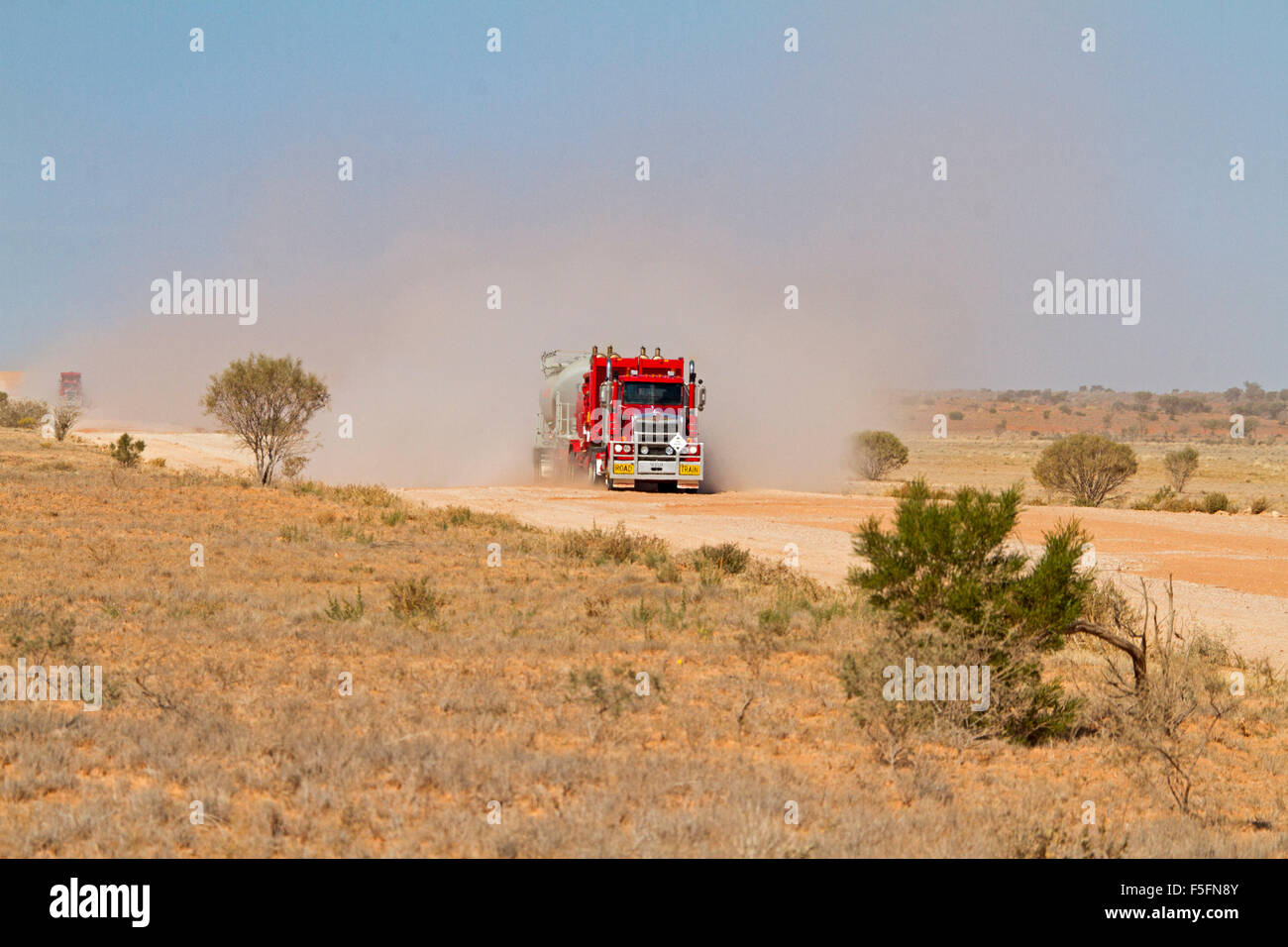 Road train, huge red semi-trailer / truck loaded with mining machinery ahead of cloud of dust on Australian outback road Stock Photo