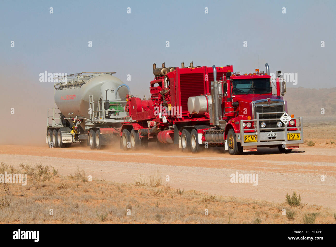 Road train, huge red semi-trailer / truck loaded with mining machinery ahead of cloud of dust on Australian outback road Stock Photo