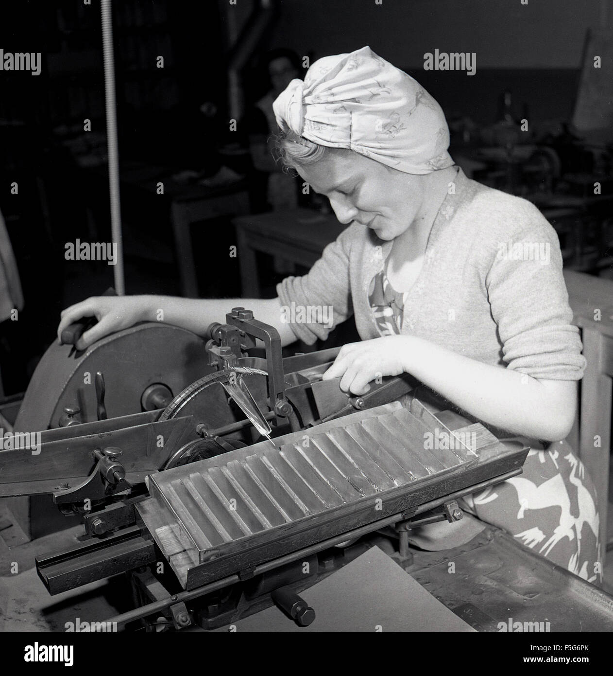 1950s historical, female operative wearing headscarf checking on industrial machine for making precision hardened steel dowel pins. Stock Photo