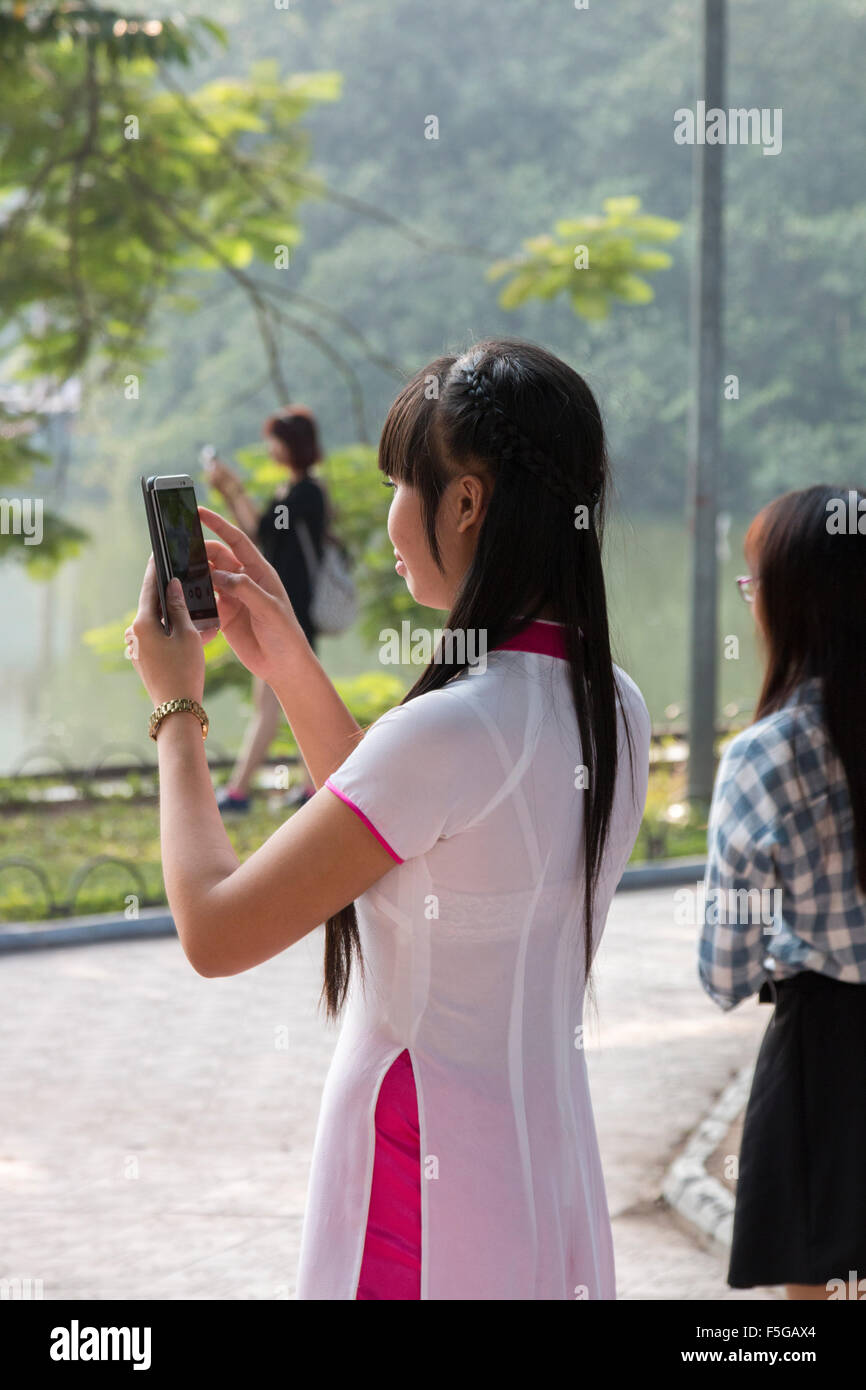 vietnamese girl lady in traditional dress ao dai, looking at the screen on a smart phone, stood by hoan kiem lake,Hanoi,Vietnam Stock Photo