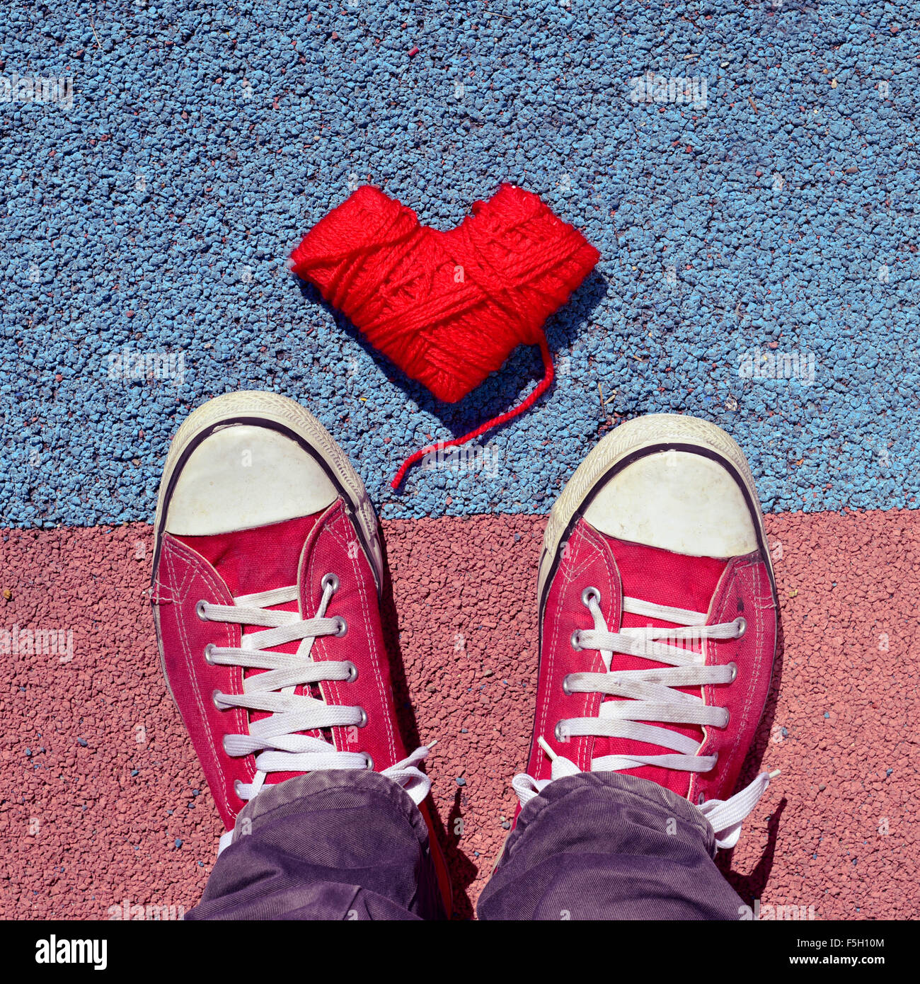 high-angle shot of a heart-shaped coil of red yarn and the feet of a man wearing red sneakers stepping on the asphalt Stock Photo