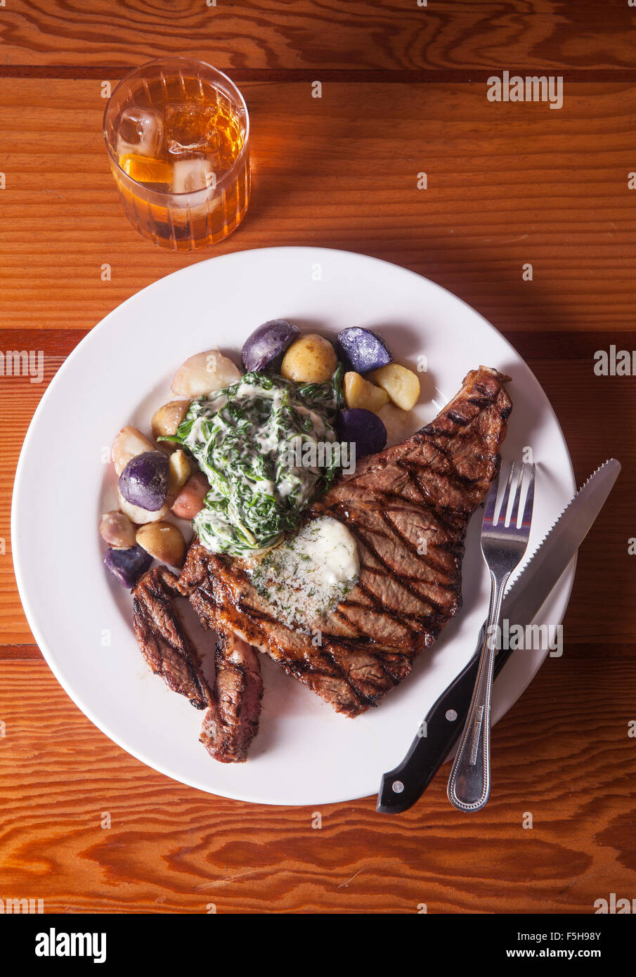 overhead view of steak with potatoes and spinach knife and fork and cocktail on wood table Stock Photo