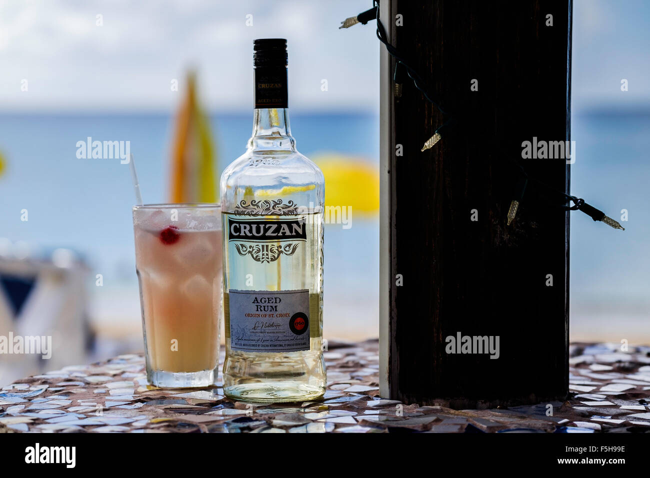 A bottle of Cruzan aged rum and a rum cocktail in a glass with a beach umbrella and Caribbean sea in background. Stock Photo