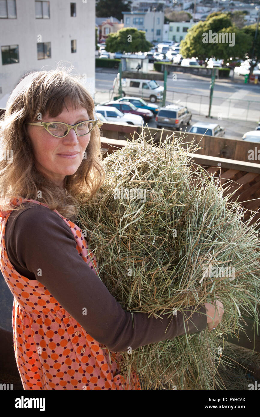 A young woman with cat-eye glasses turns back to look at the camera holding straw in her San Francisco urban farm Stock Photo