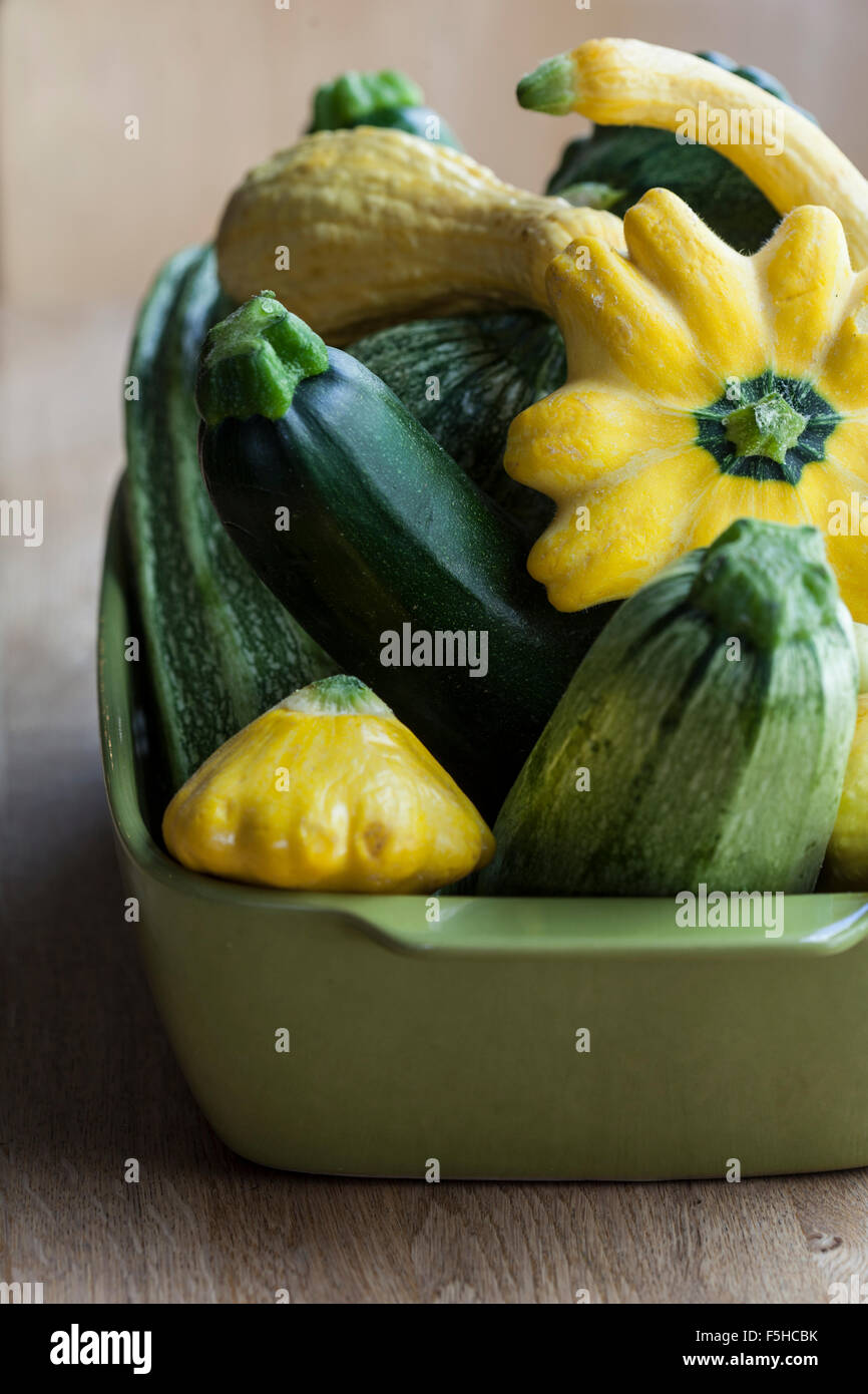 a square green container holds a variety of summer squash on a wood table Stock Photo