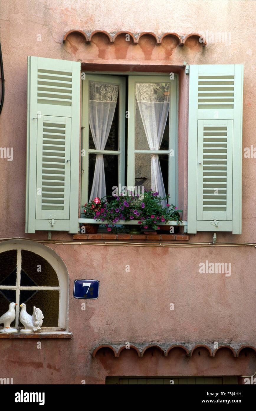 Exterior of a pink French cottage with pale green shutters on a window with lace curtains Stock Photo