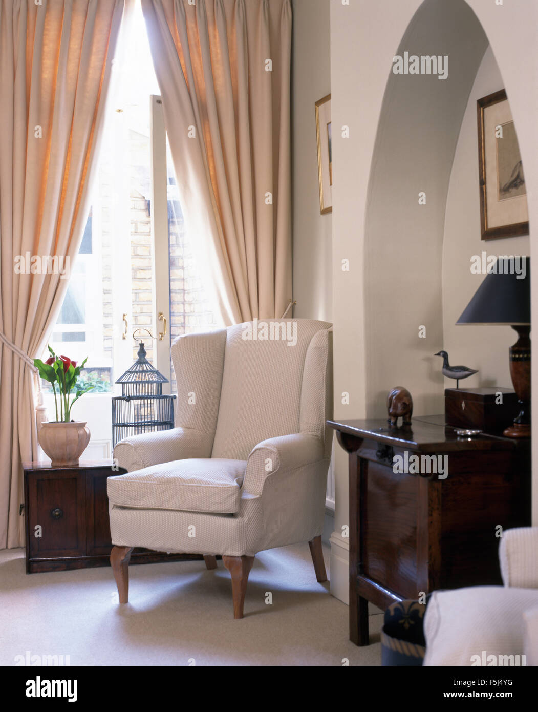 White wing chair in front of window with cream drapes in townhouse living room with an antique chest in the alcove Stock Photo