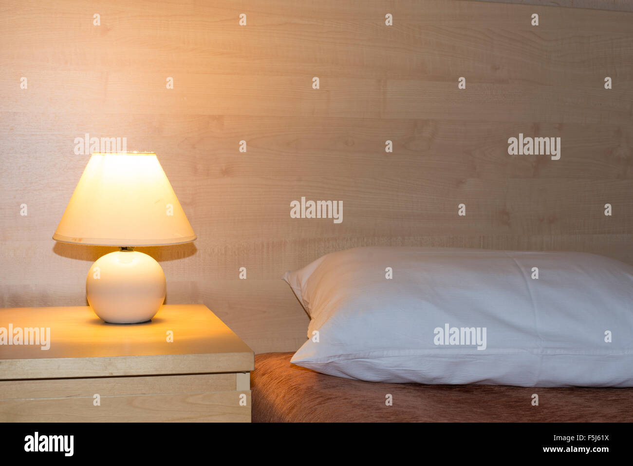 simple room interior with illuminated bed light on wooden table and focus on pillow Stock Photo