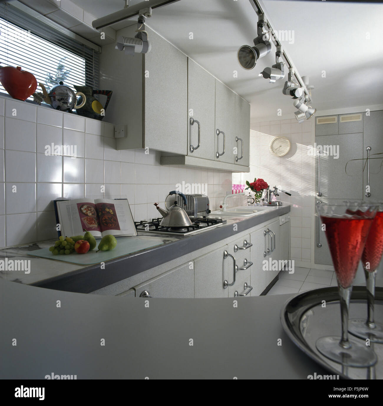 Track spotlights and white tiling in a nineties kitchen with pale gray fitted units and an Alessi kettle on the hob Stock Photo