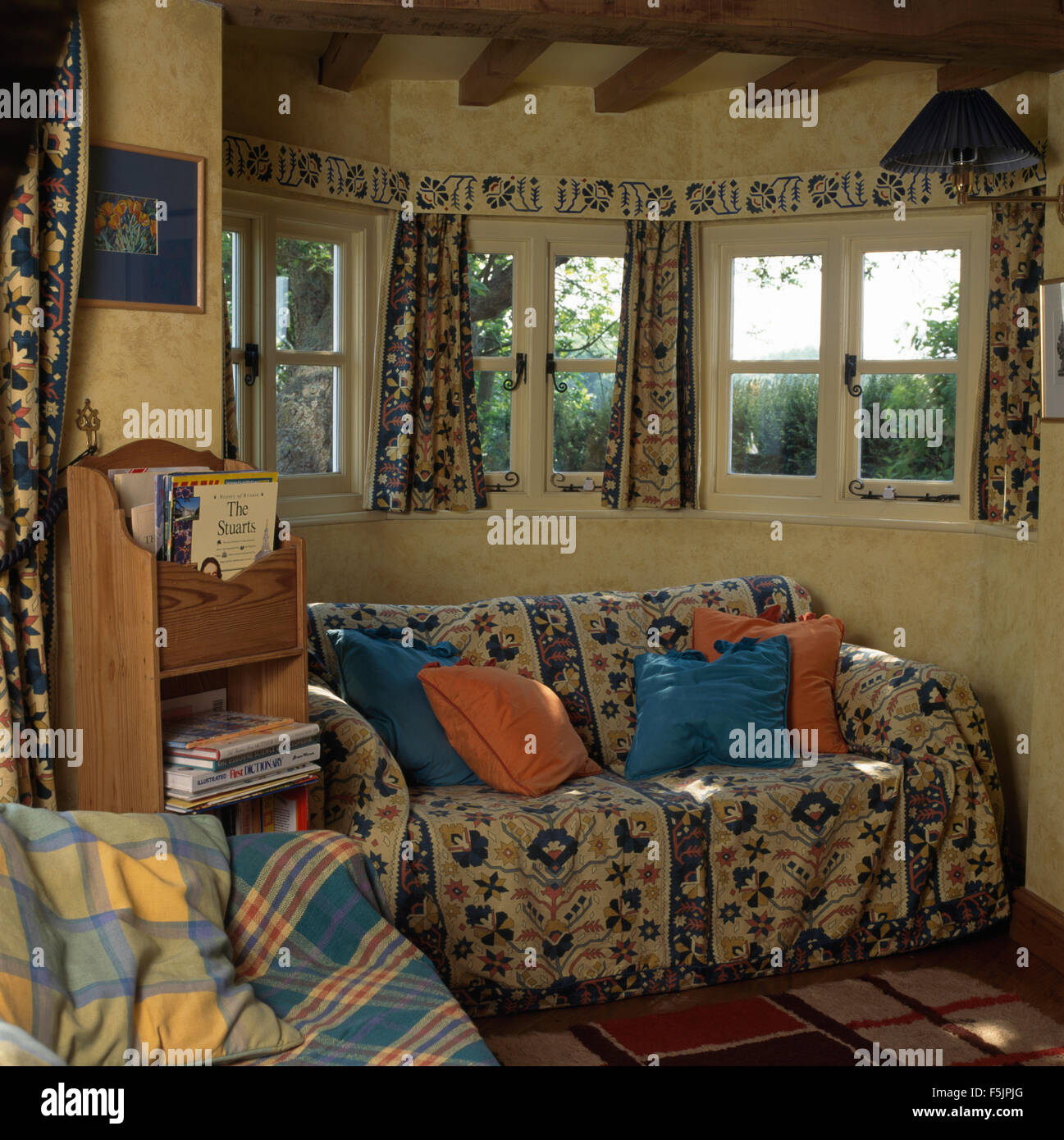 Floral patterned throw on sofa below windows in economy style cottage living room Stock Photo